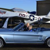 Two people sitting in a blue 1965 classic Ford Mustang convertible, parked in front of a silver and red P-51 Mustang fighter plane.