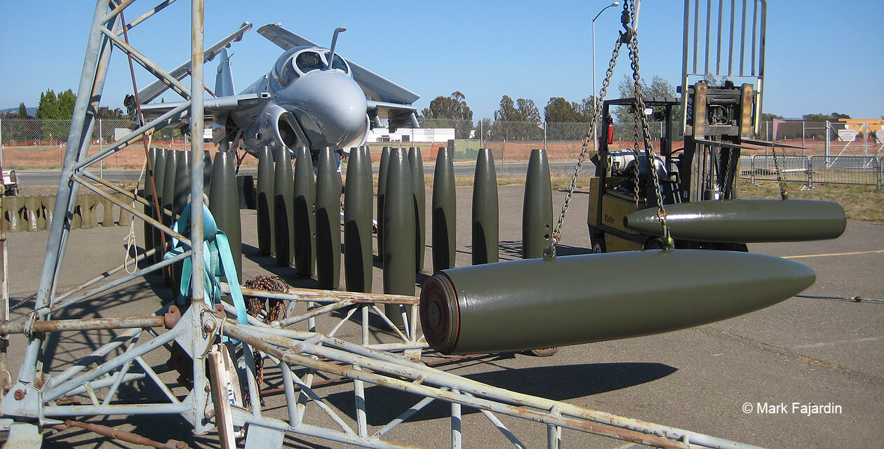 a6e-intruder-pcam-05 Several freshly painted bombs without tail assemblies waiting to be loaded onto an A-6E Intruder attack jet that sits in the background. One bomb hangs suspended from a small crane.