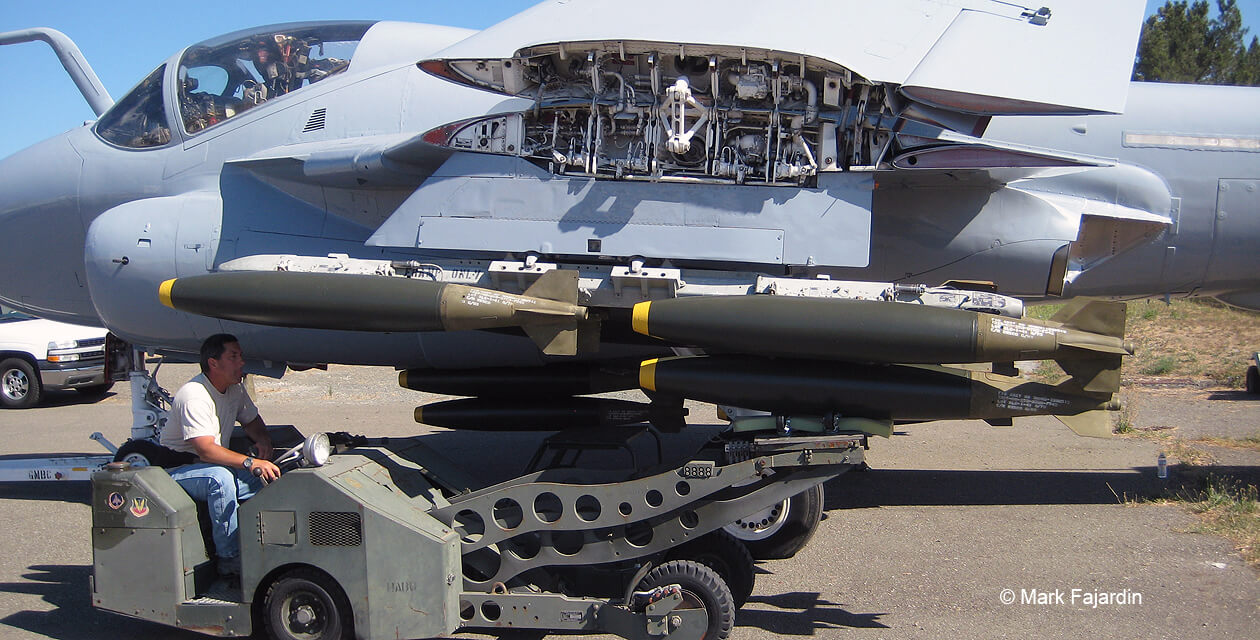 a6e-intruder-pcam-07 A man on an ordnance loading tractor lifts a cluster of inert bombs onto the underwing pylon of an A-6E Intruder.