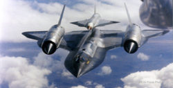 In-flight photo of M-21 mother ship which looks like an SR-71 Blackbird, carrying a D-21 drone on its back. View is from the front and above, and a layer of clouds can be seen below.