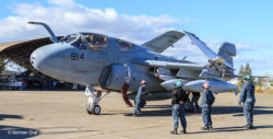 EA-6B Prowler electronic warfare jet aircraft on the concrete ramp in front of a curved roof hangar. Ground crew scramble around preparing the plane for engine shut-down.