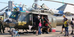 UH-1H Huey helicopter parked as a static display aircraft at an air show, with other planes parked around it and visitors climbing aboard.